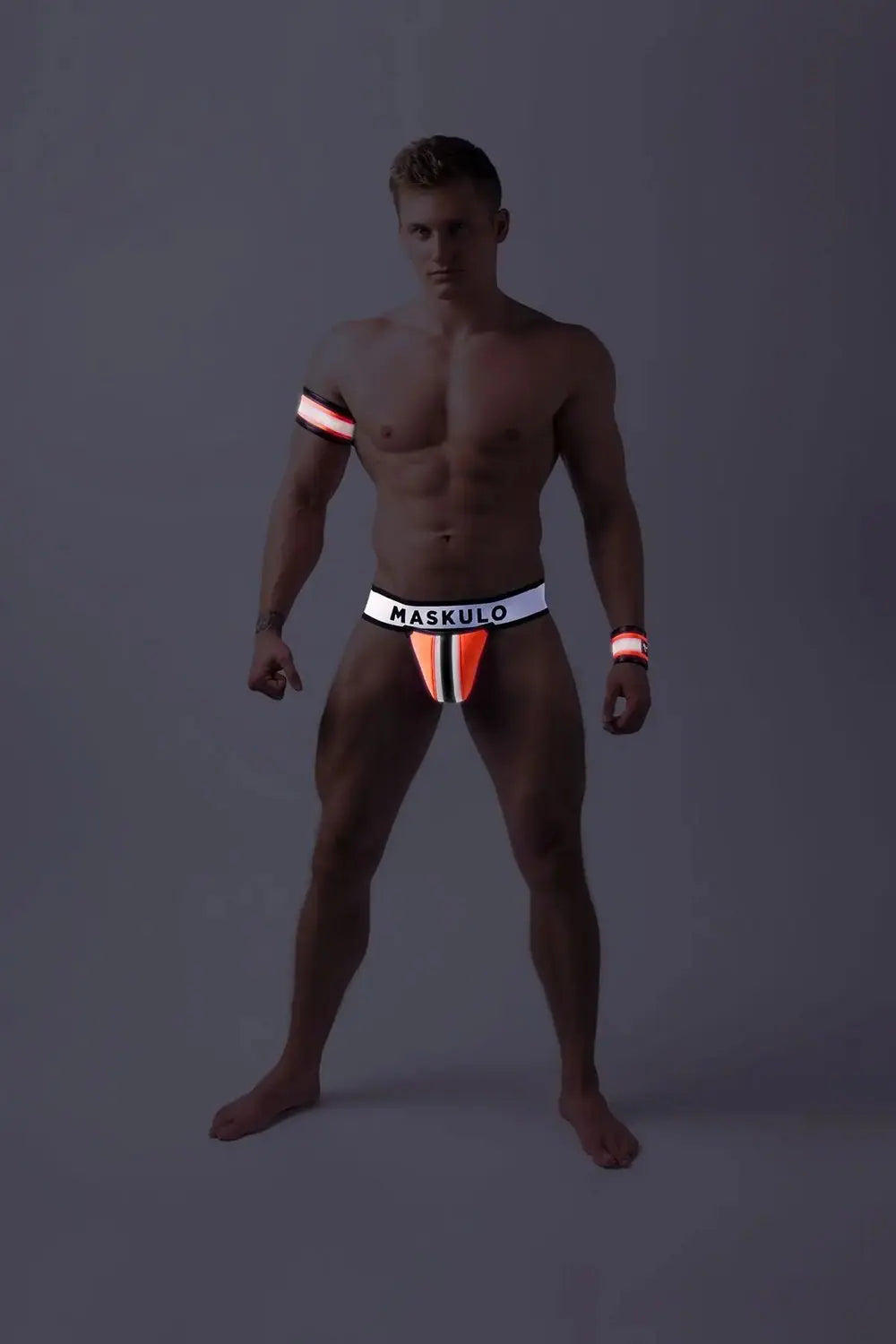 Maskulo Youngero neon orange fetish jockstrap for men with detachable codpiece, UV glowing accents, and black shiny trim, ideal for urban Gen Z and gender-fluid style